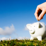 Person's hand putting coin in a piggy bank that is sitting on a grassy hill