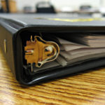 A close-up, side view of a black three-ring binder, filled with papers.