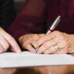Estate planning attorney assisting elderly woman holding a pen with signing estate planning documents.